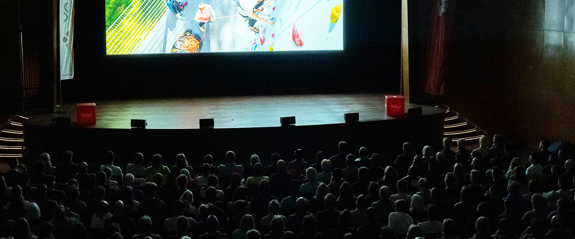 Darkened Banff Tour venue with a full audience in front of a large screen showing a climbing scene on an outdoor climbing wall.