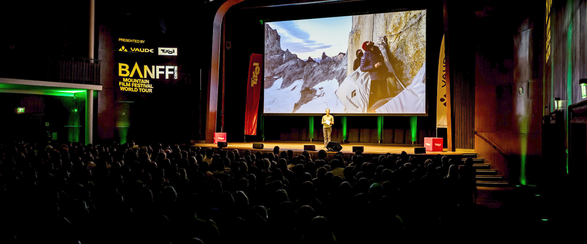 Host on stage at a Banff Tour event in front of a large screen showing two climbers in a portaledge on a rock face.
