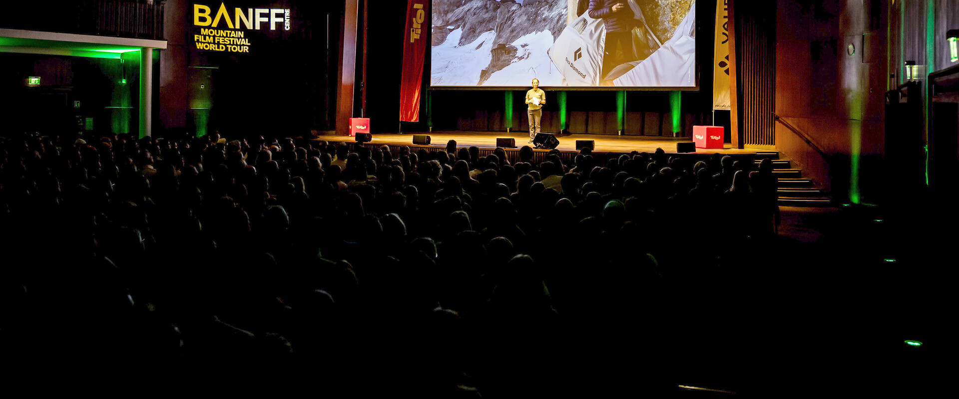 Moderator auf der B&uuml;hne eines Banff Tour Events vor einer gro&szlig;en Leinwand, die zwei Bergsteiger:innen in einem Portaledge an einer Felswand zeigt.