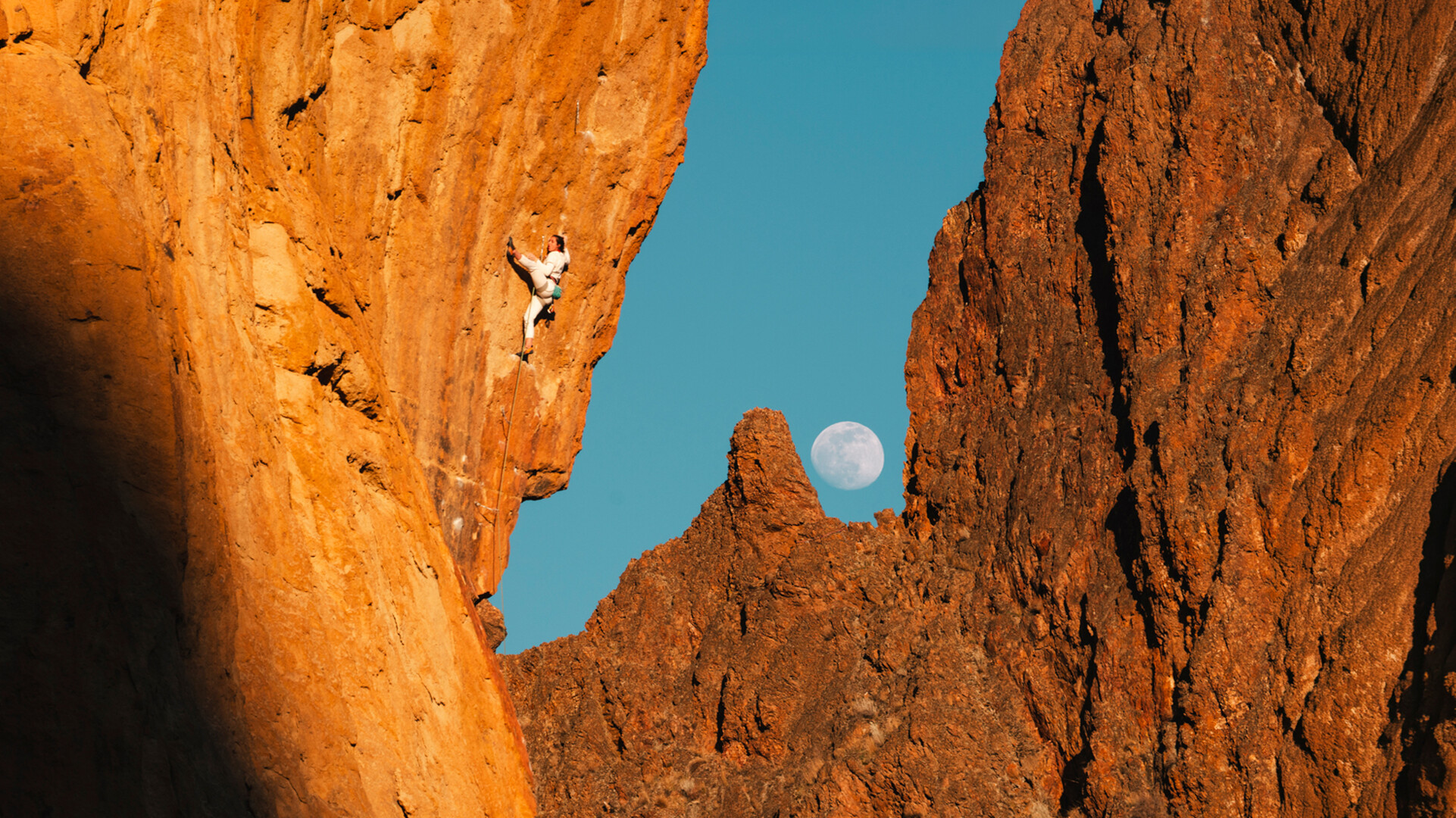 Climber on a steep, overhanging rock face glowing orange, with a full moon rising over jagged rock spires against a blue sky in the background.