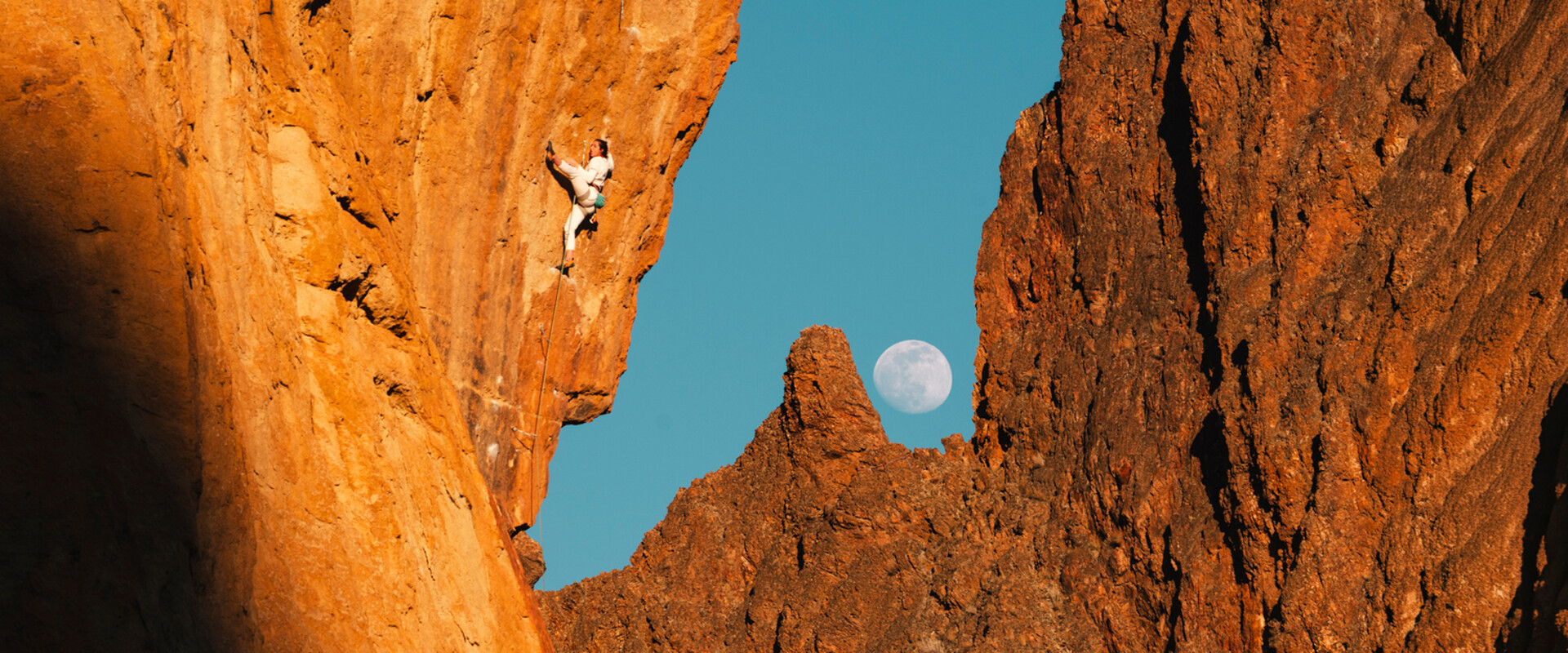 Climber on a steep, overhanging rock face glowing orange, with a full moon rising over jagged rock spires against a blue sky in the background.