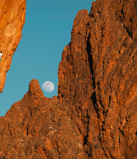 Climber on a steep, overhanging rock face glowing orange, with a full moon rising over jagged rock spires against a blue sky in the background.