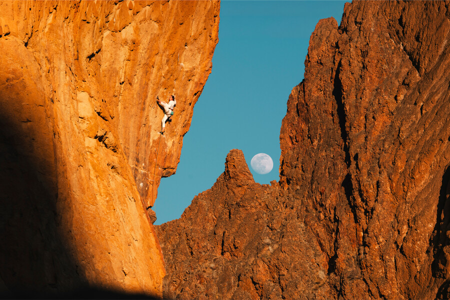 Climber on a steep, overhanging rock face glowing orange, with a full moon rising over jagged rock spires against a blue sky in the background.