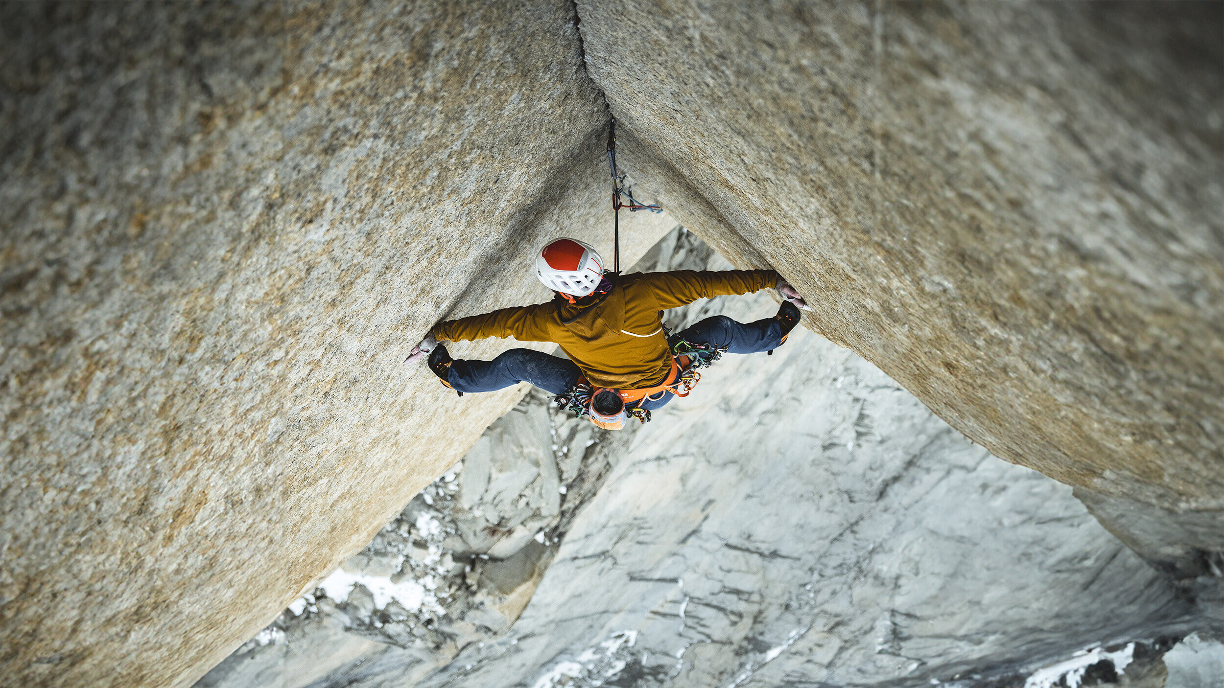 Climber Siebe Vanhee wedged in a crack of an overhanging granite wall on the route Riders on the Storm in Patagonia, shot from above, with deeply snow-covered rock in the background.