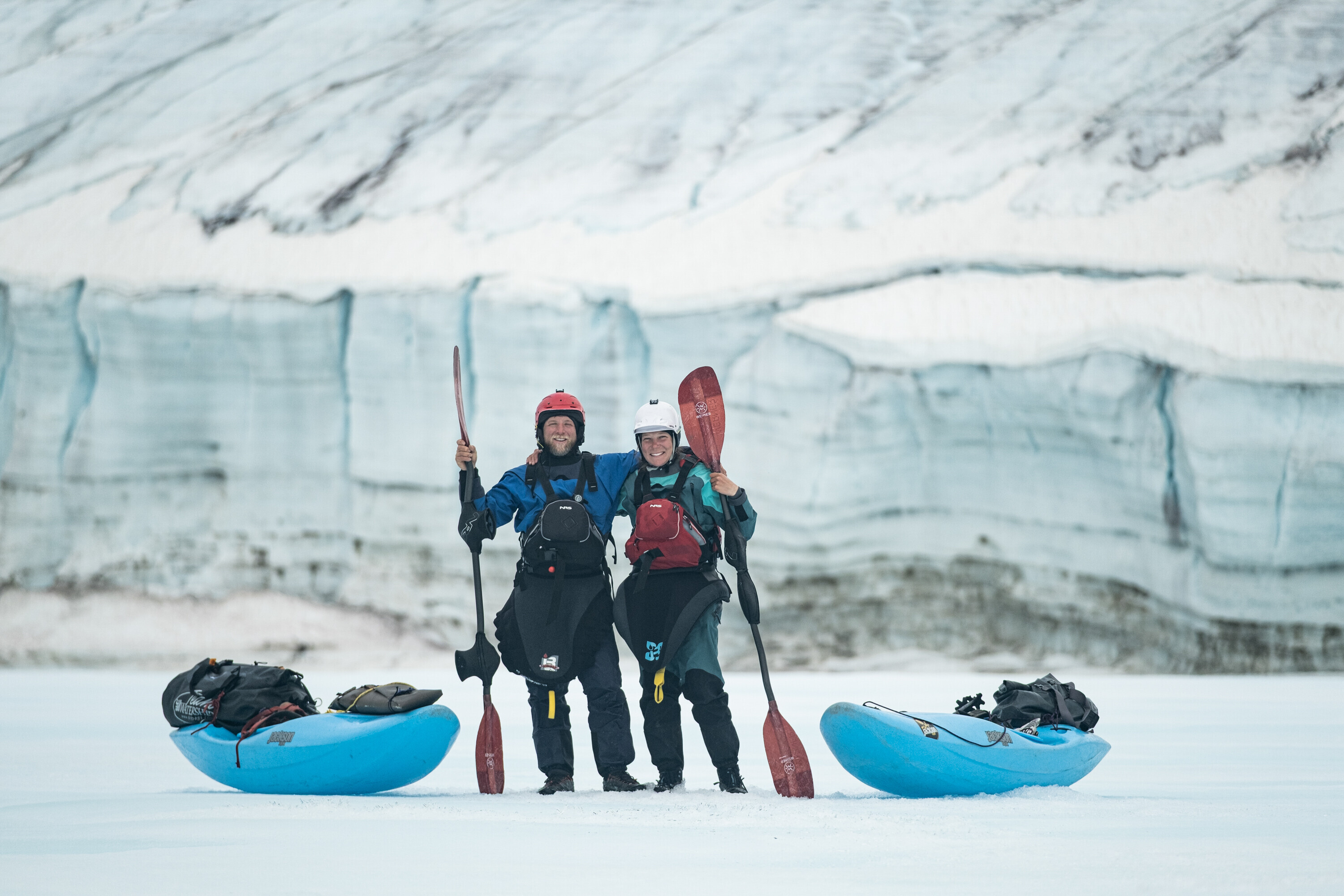 Two kayakers, Sarah McNair-Landry and Erik Boomer, in drysuits holding paddles, smiling between two blue kayaks on a glacier in front of an ice wall.