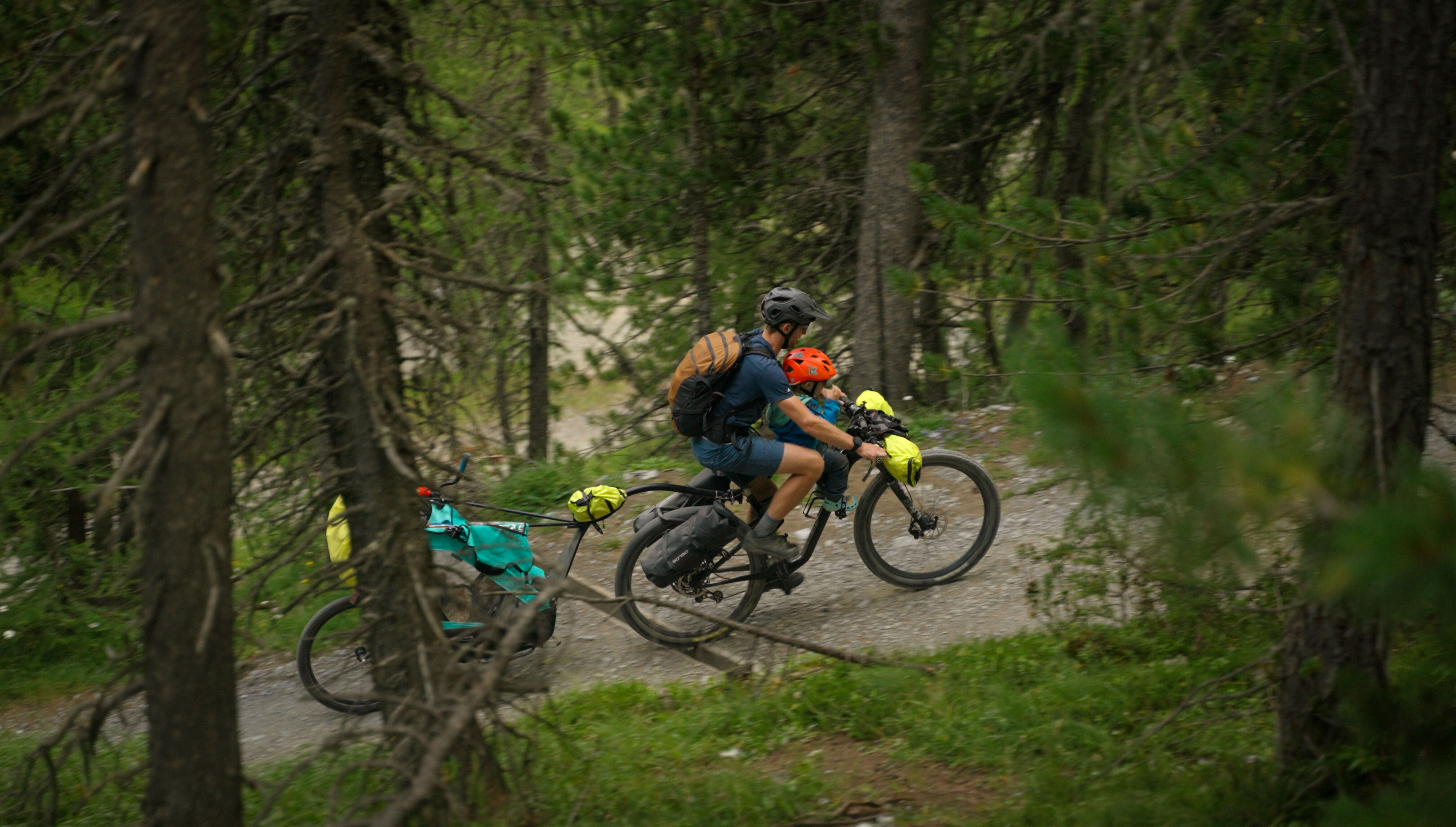An adult and a child riding together on a loaded mountain bike with a trailer along a forest path.